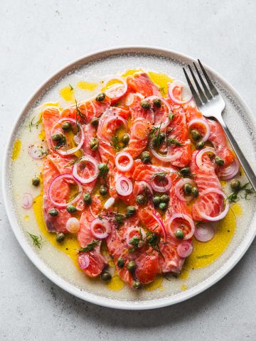 Salmon Crudo served on a plate - slices of sashimi grade raw salmon topped with a lemon olive oil dressing, onion, cxapers and dill.